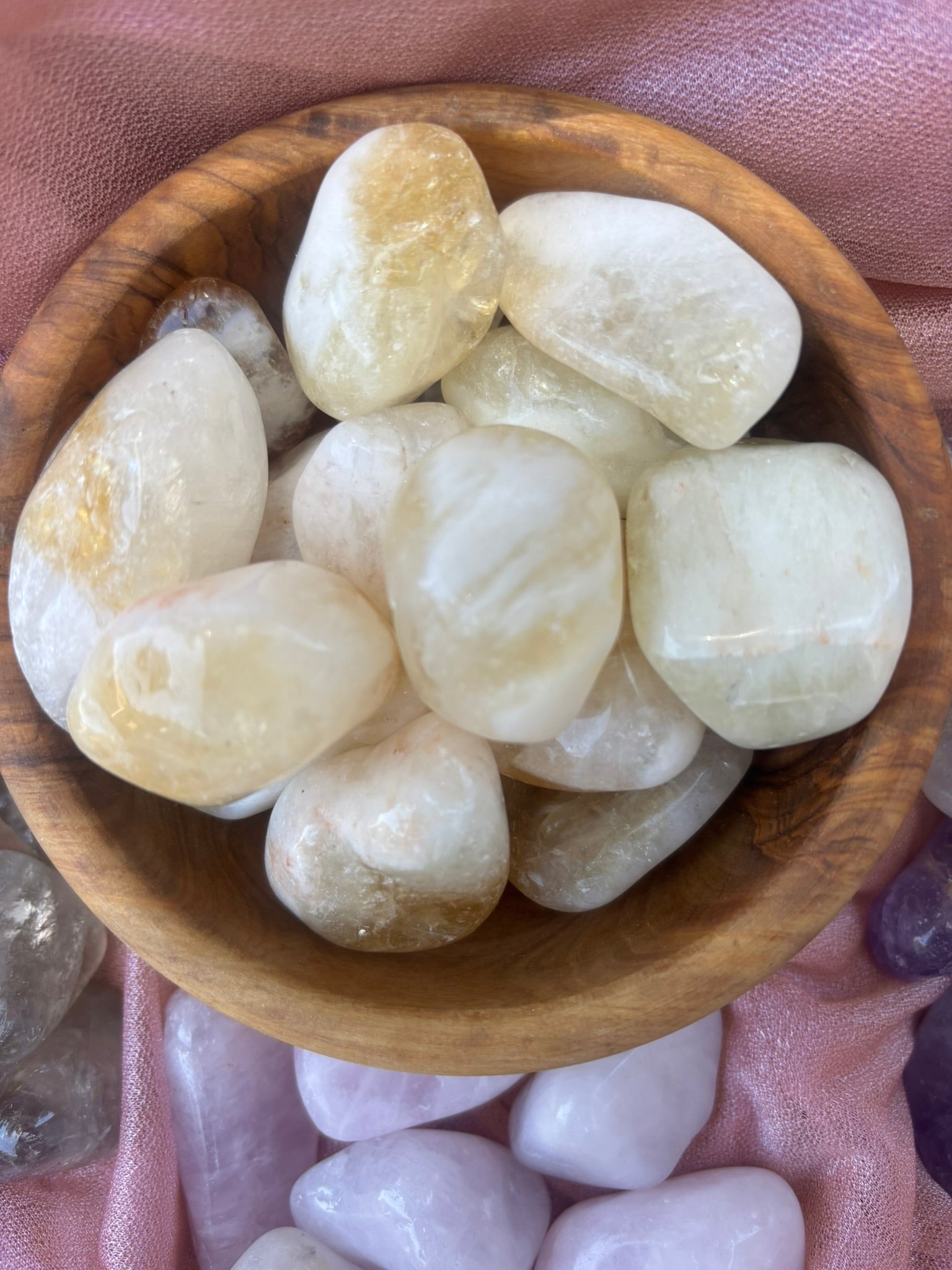 A wooden bowl filled with smooth, polished tumbled stones in soft cream, pale yellow, and translucent tones, set against pink fabric with additional stones around the edges.