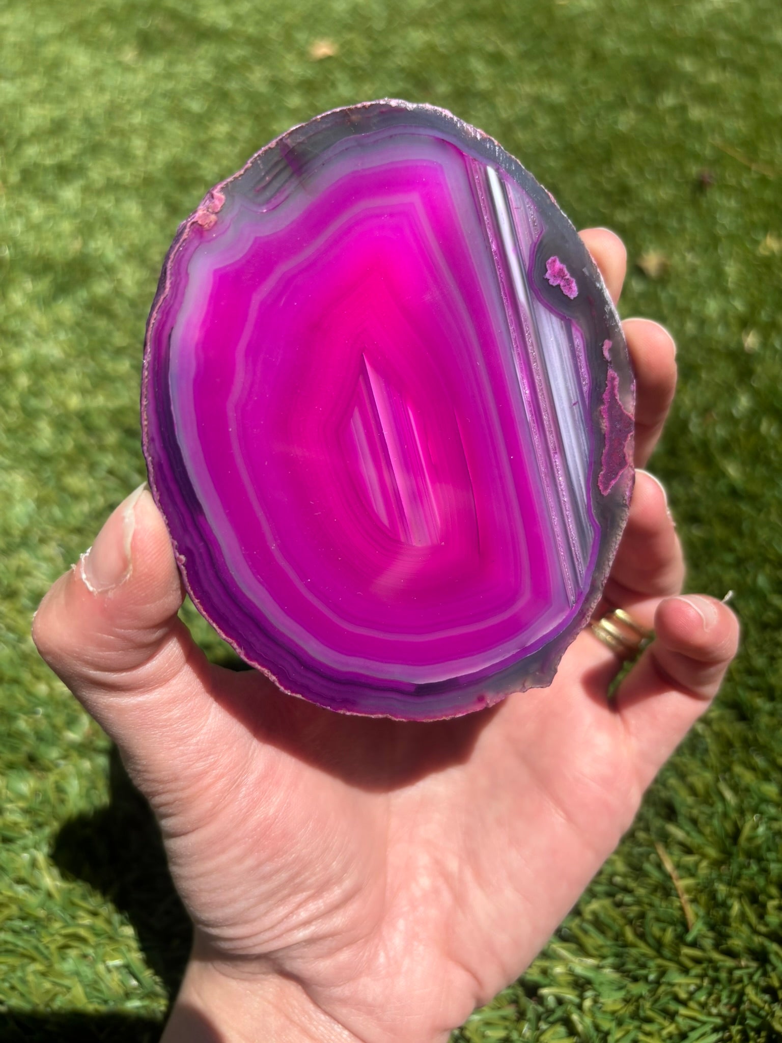 A hand holding a polished slice of magenta and purple agate stone against a background of green grass.